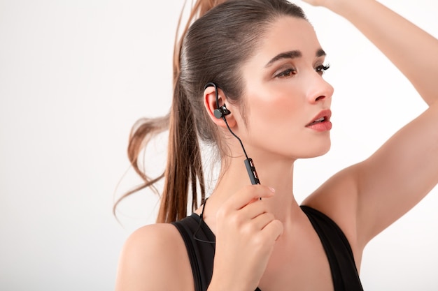 Atractiva mujer morena en jogging top negro escuchando música en auriculares posando aislado en la pared blanca