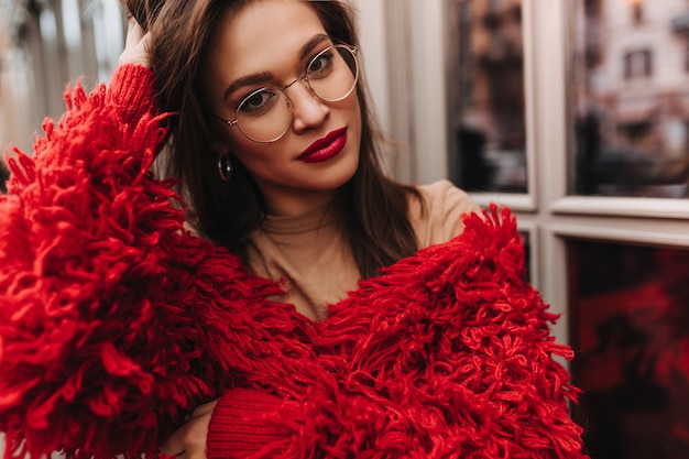 Atractiva mujer morena de ojos marrones con lápiz labial rojo vestida con chaqueta de punto brillante y top beige mirando a cámara a través de gafas contra ventanas.