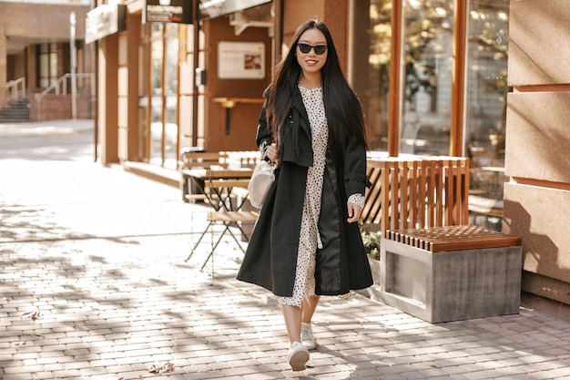 Atractiva mujer morena de pelo largo con gabardina negra y vestido midi sonríe sinceramente y camina afuera