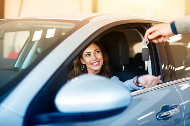 Una atractiva mujer morena sentada en su coche nuevo y tomando las llaves del concesionario de vehículos