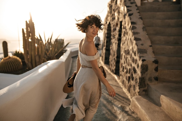 Atractiva mujer morena con vestido beige sostiene sombrero de paja y corre a lo largo del camino en la hermosa ciudad vieja Niña feliz sonríe y mira a la cámara