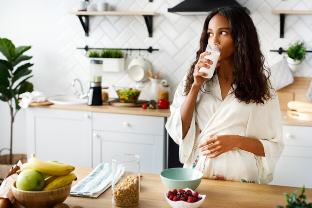 La atractiva mujer mulata sonríe está bebiendo leche cerca de la mesa con frutas frescas en la moderna cocina blanca vestida con ropa de dormir con el pelo suelto y mirando a la derecha