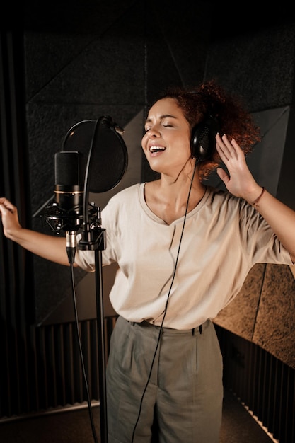 Atractiva mujer músico en auriculares cantando emocionalmente en estudio de grabación de sonido