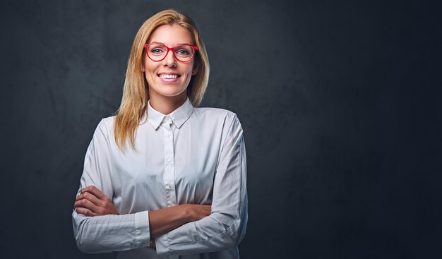 Atractiva mujer de negocios rubia con camisa blanca, anteojos y brazos cruzados sobre fondo gris.