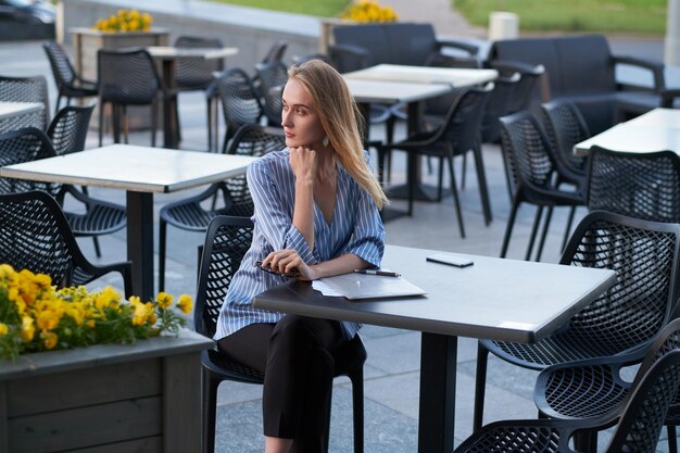 Atractiva mujer de negocios rubia está pensando en su trabajo mientras está sentada afuera de la cafetería con papeles.