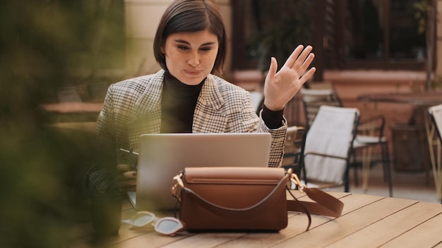 Atractiva mujer de negocios saludando hablando con colegas por videollamada en la computadora portátil durante el descanso para tomar café en la cafetería al aire libre Expresión de saludo