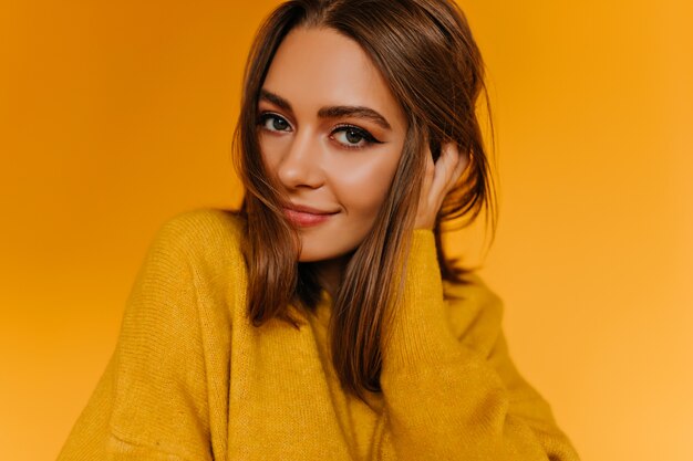 Atractiva mujer de ojos marrones jugando con su cabello. Impresionante chica blanca en suéter sonriendo suavemente en la pared naranja.