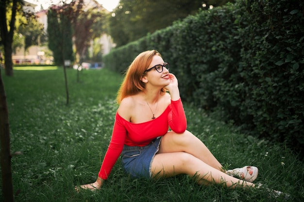 Atractiva mujer pelirroja con anteojos en blusa roja y falda de jeans posando en Green Park