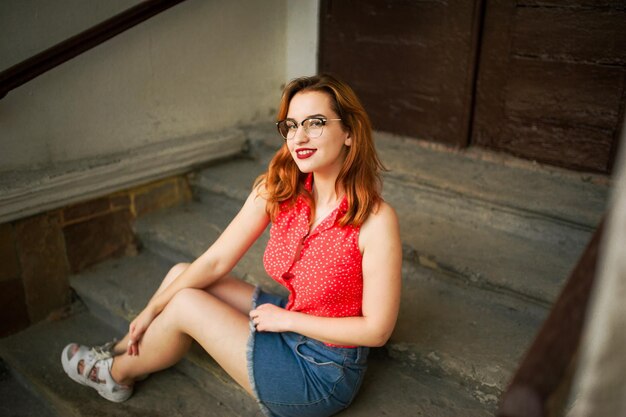 Atractiva mujer pelirroja con anteojos en blusa roja y falda de jeans posando