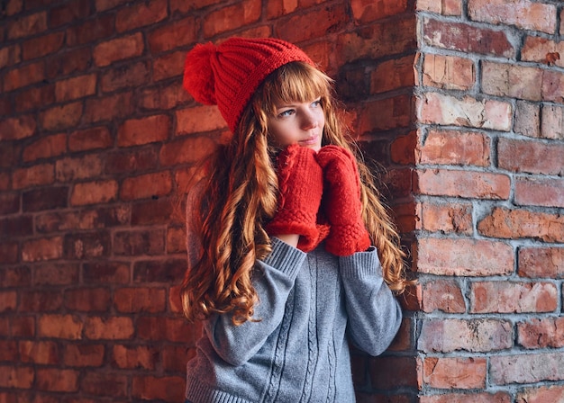 Una atractiva mujer pelirroja vestida con un gorro cálido y guantes posando sobre la pared de un ladrillo rojo.