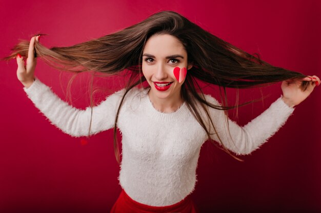 Atractiva mujer de pelo castaño en suéter blanco disfrutando de la sesión de fotos en el día de San Valentín