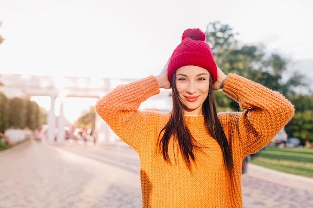 Atractiva mujer de pelo castaño en suéter naranja de moda sonriendo en la ciudad
