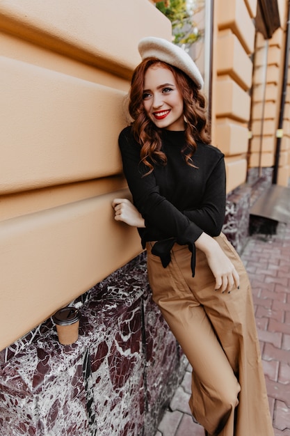 Atractiva mujer de pelo largo con pantalones marrones de pie en la calle. Foto al aire libre de una chica francesa de buen humor.