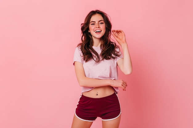 Atractiva mujer de pelo oscuro rizado en camiseta y pantalones cortos está sonriendo en la pared rosa
