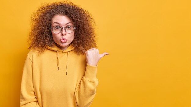 Atractiva mujer de pelo rizado sorprendida mantiene los labios redondeados con el pulgar hacia fuera hace su elección en las compras vestida con poses casuales con capucha sobre la pared amarilla muestra el logotipo o la oferta promocional en el espacio de la copia