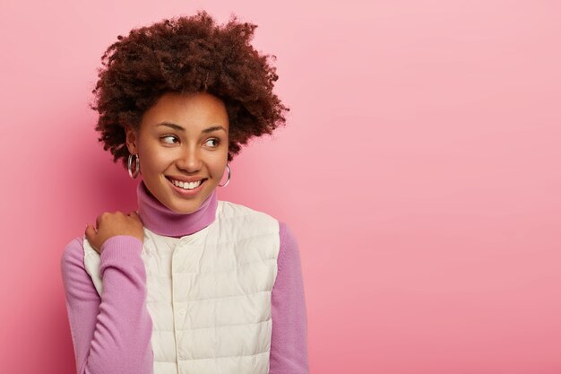 Atractiva mujer de pelo rizado toca el hombro, usa cuello alto y chaleco blanco, está de buen humor