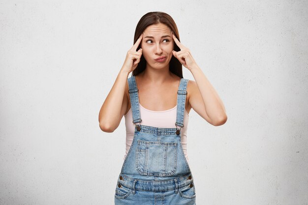 Atractiva mujer pensativa vestida casualmente, manteniendo los dedos en las sienes, curvando los labios y mirando a un lado pensando en algo importante, tratando de concentrarse, aislado