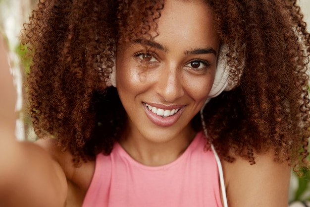 Atractiva mujer de piel oscura con cabello oscuro rizado y tupido, usa audífonos, escucha audiolibros, muestra una sonrisa agradable, hace selfie, disfruta del tiempo libre. Estudiante descansa después de las lecciones universitarias