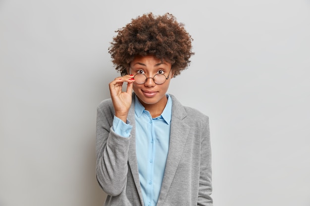 Atractiva mujer de piel oscura tiene la mano en las gafas se ve agradablemente tiene el pelo rizado