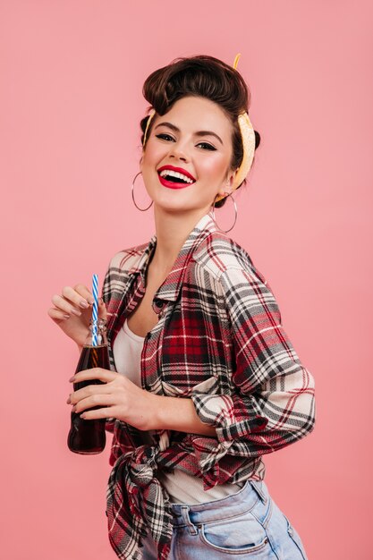 Atractiva mujer pinup en camisa a cuadros sonriendo a la cámara. Disparo de estudio de una atractiva chica morena aislada sobre fondo rosa con botella.