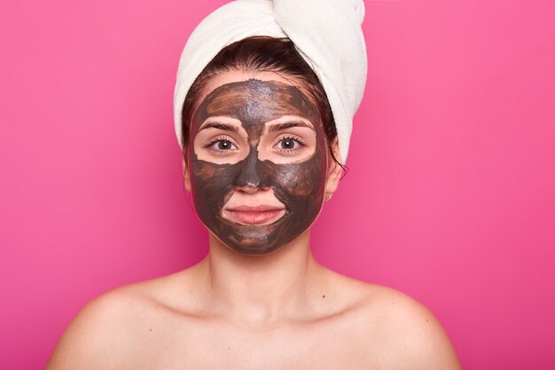 Atractiva mujer posa con expresión facial seria y tranquila, tiene máscara de chocolate en la cara, con hombros desnudos, cuida su belleza y apariencia, usa una toalla blanca en la cabeza. Concepto de cuidado de la piel.