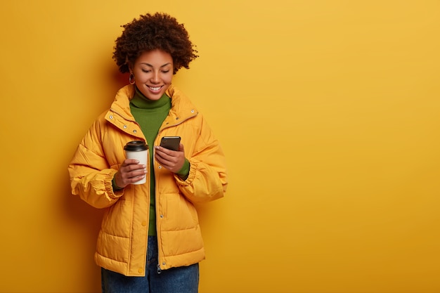 Atractiva mujer positiva en ropa de abrigo amarilla, feliz de leer buenos comentarios debajo de la publicación, sostiene un teléfono celular moderno, bebe café para llevar
