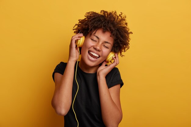 Atractiva mujer rizada con grandes sonrisas, mantiene las manos en el auricular, vestida con camiseta negra, aislada sobre fondo amarillo