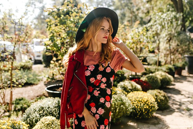 Atractiva mujer rizada con sombrero de ala ancha, chaqueta de cuero roja y vestido negro con estampado floral mira pensativamente a la distancia, disfrutando del día de primavera en el jardín.