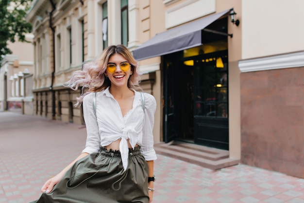 Atractiva mujer rizada con sonrisa sincera jugando con su falda larga mientras camina por la calle