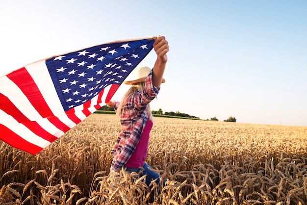 Atractiva mujer rubia caminando por el campo de trigo y sosteniendo la bandera ondeante de Estados Unidos