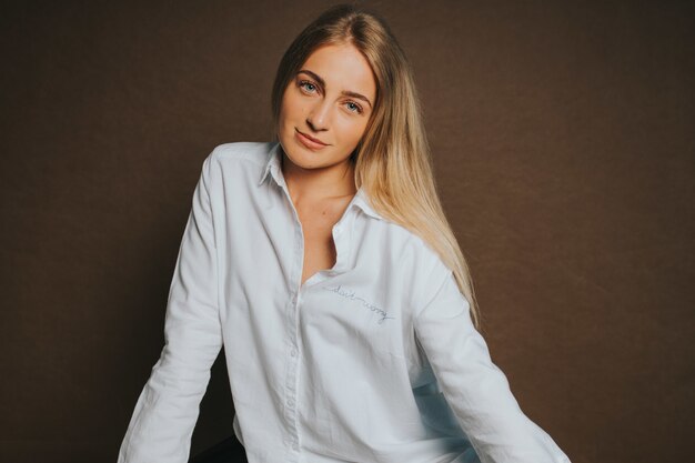 Atractiva mujer rubia caucásica con una camisa blanca posando en una pared marrón