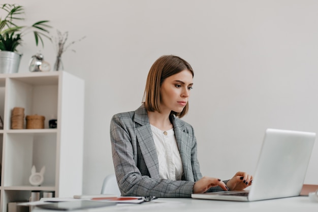 Atractiva mujer rubia escribiendo carta en la computadora portátil en su lugar de trabajo. retrato de dama de elegante chaqueta en oficina brillante.