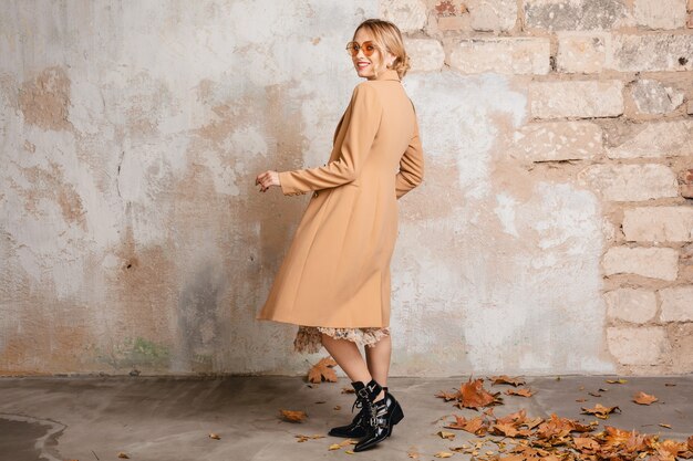 Atractiva mujer rubia con estilo en abrigo beige caminando en la calle contra la pared vintage