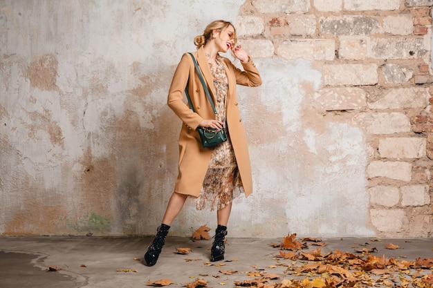 Foto gratuita atractiva mujer rubia con estilo en abrigo beige caminando en la calle contra la pared vintage