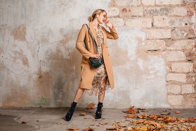 Atractiva mujer rubia con estilo en abrigo beige caminando en la calle contra la pared vintage