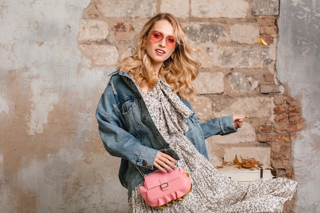 Atractiva mujer rubia con estilo en jeans y chaqueta de gran tamaño caminando contra la pared en la calle