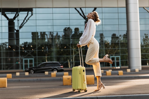 Atractiva mujer rubia levanta coquetamente la pierna y posa con equipaje Feliz hermosa chica en blusa blanca y pantalones beige sostiene maleta cerca del aeropuerto