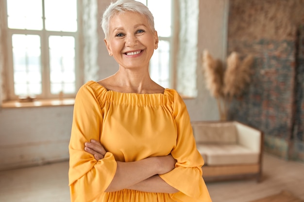 Atractiva mujer rubia madura segura con vestido amarillo cruzando los brazos sobre el pecho y sonriendo felizmente a la cámara, posando en su apartamento recientemente renovado con sofá y ventanas en segundo plano.