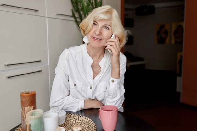 Atractiva mujer rubia de mediana edad descansando en su casa en la cocina y hablando por teléfono móvil