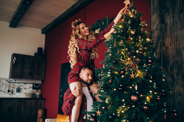 Atractiva mujer rubia en pijama rojo poniendo estrella de Navidad en el árbol de Navidad. Vacaciones juntos.