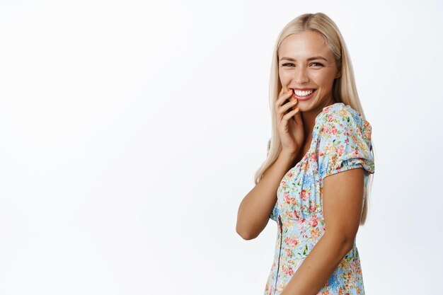 Atractiva mujer rubia riendo sonriendo coqueta mirando coqueta a la cámara posando en lindo vestido de verano sobre fondo blanco.