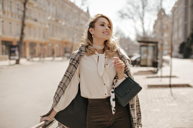 Atractiva mujer rubia rizada con abrigo de tweed a cuadros, blusa blanca y pantalones marrones sonríe sinceramente sostiene el bolso y camina al aire libre