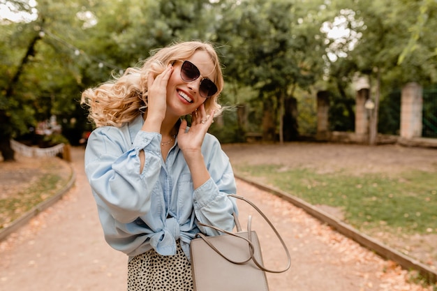 Atractiva mujer rubia sincera caminando en el parque con elegante traje con elegantes gafas de sol y bolso