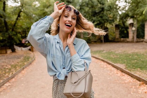 Atractiva mujer rubia sincera caminando en el parque con elegante traje con elegantes gafas de sol y bolso