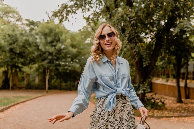 Atractiva mujer rubia sonriente caminando en el parque en traje elegante