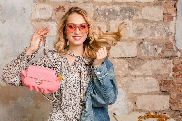 Atractiva mujer rubia sonriente elegante en chaqueta de jeans caminando contra la pared en la calle