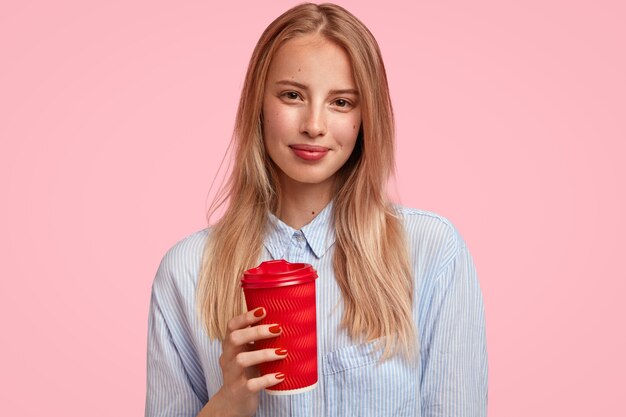 Atractiva mujer rubia sostiene una bebida caliente en un vaso de papel desechable, viste una camisa elegante, se para contra la pared rosa, se rompe después de dar conferencias. Concepto de personas y tiempo libre.