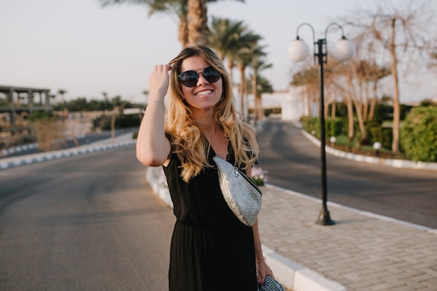 Atractiva mujer rubia en vestido negro y elegantes gafas de sol posando al aire libre con exóticas palmeras. Retrato de mujer joven con lindo peinado y bolso plateado caminando por la calle