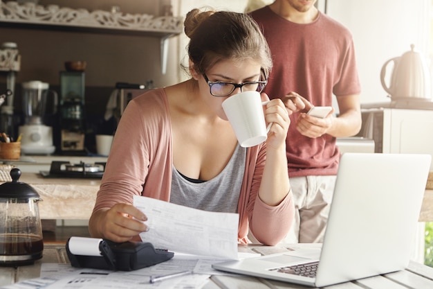 Atractiva mujer seria en espectáculos tomando café y estudiando documentos en sus manos, administrando el presupuesto familiar y haciendo trámites en la mesa de la cocina con pila de facturas, computadora portátil y calculadora