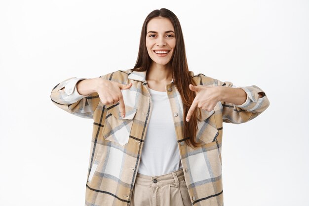 Atractiva mujer sonriente apuntando con el dedo hacia la pancarta, mostrando el logotipo u oferta promocional, recomendando visitar la tienda, de pie sobre una pared blanca